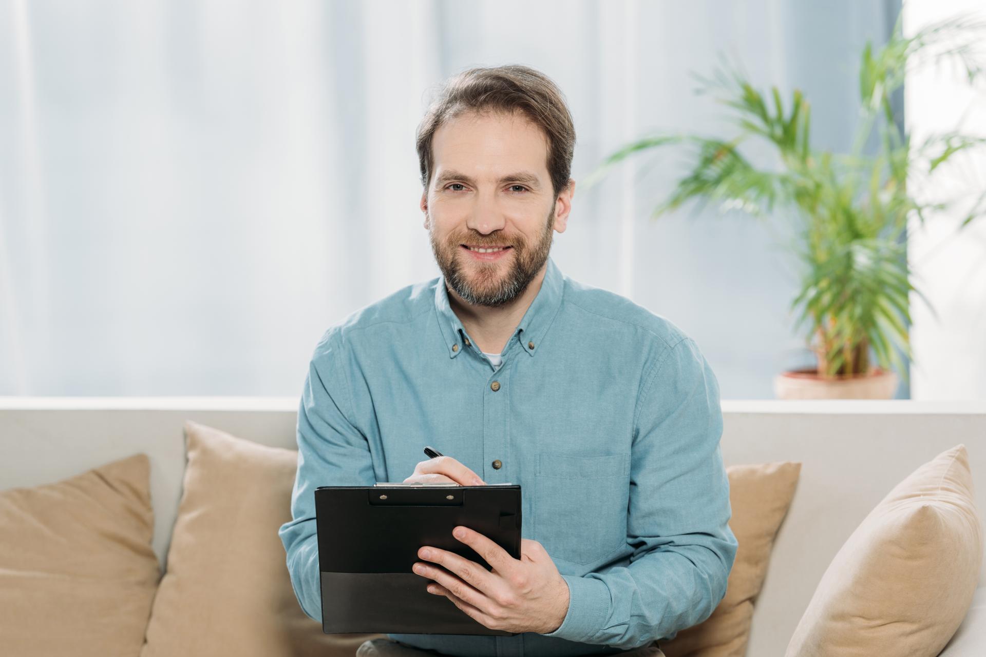 bearded male psychotherapist writing on clipboard and smiling at camera